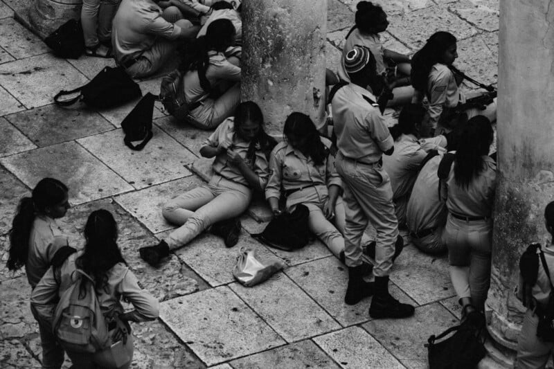 A group​ of people dressed in uniform⁢ relax on a‍ stone pavement, some ‍leaning against pillars. They appear​ to be resting, ⁤with several sitting and one⁤ person standing. The scene is captured in black and white.