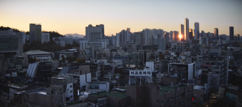 Panoramic view of a city skyline at sunset, featuring a mix of high-rise buildings and smaller structures. The sun reflects off one of the taller buildings, casting a warm glow over the urban landscape.