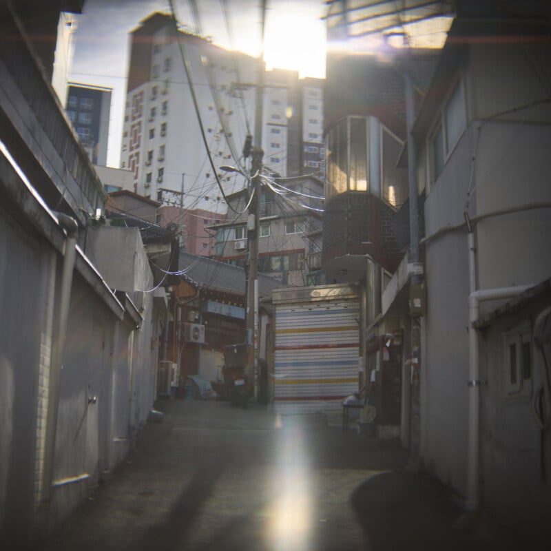 Narrow alleyway with overhead power lines and a distant view of tall buildings. Sunlight creates a lens flare, highlighting the urban setting. The street is empty, with a closed shop featuring a colorful shutter on the right.