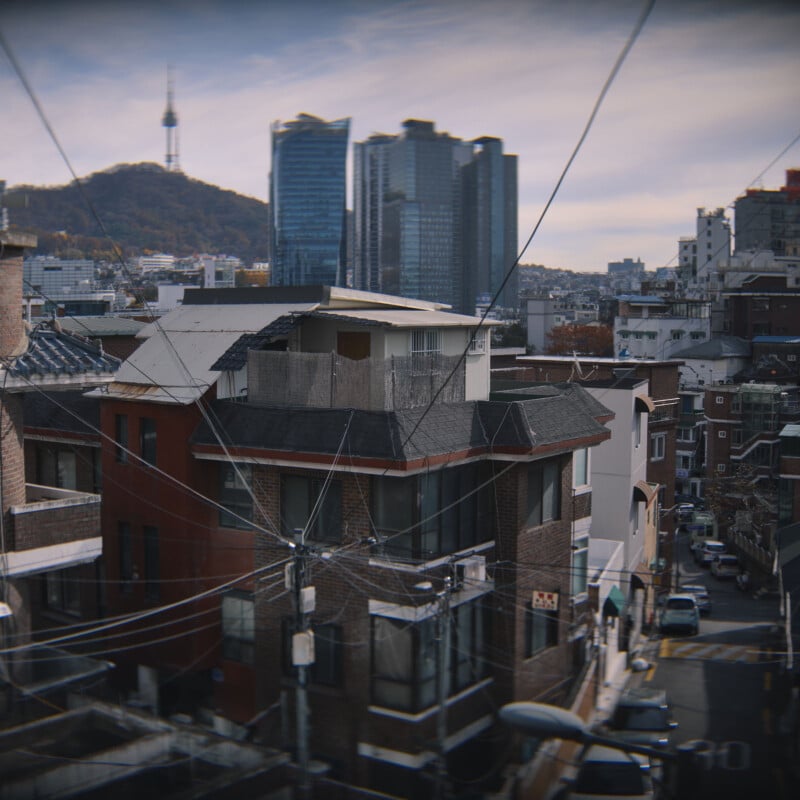 View of a cityscape with brick buildings in the foreground, intersecting power lines, and a distant tower on a hill. A cluster of modern skyscrapers stands against a partly cloudy sky.