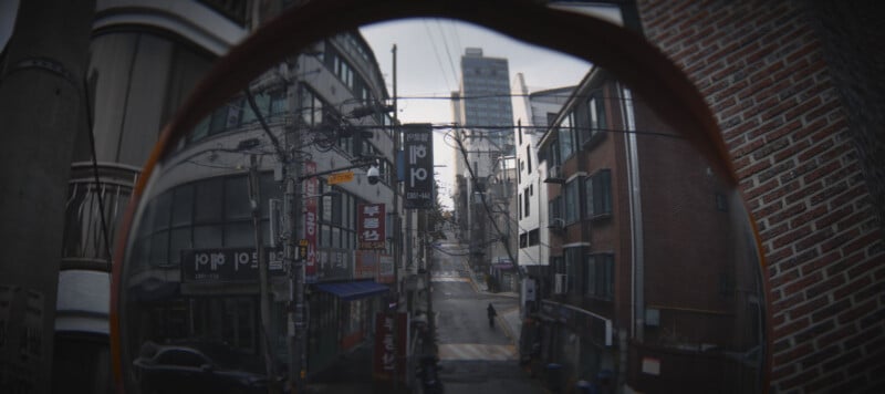 A fisheye view of an urban street scene featuring tall buildings with signs in Korean. There is a person in the distance walking along the sidewalk on a cloudy day, surrounded by power lines and various store signs.