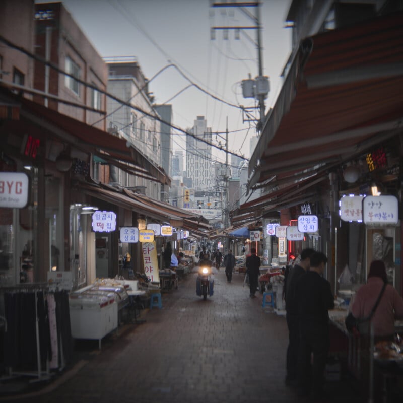 A bustling street market with overhead awnings in an urban setting. People browse outdoor stalls displaying goods, and a motorbike rides down the narrow pathway. Illuminated signs with Korean characters hang from the stalls, creating a vibrant atmosphere.