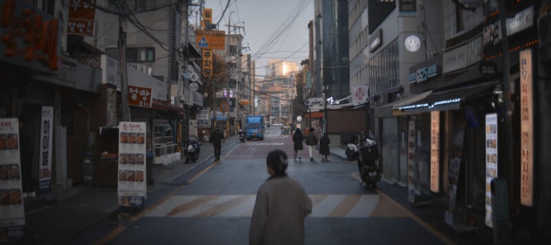 A person walks down an urban street lined with various shops and signs, including restaurants with illuminated displays. Vehicles and a blue truck are in the distance. The street is bustling with signs and electric wires overhead.