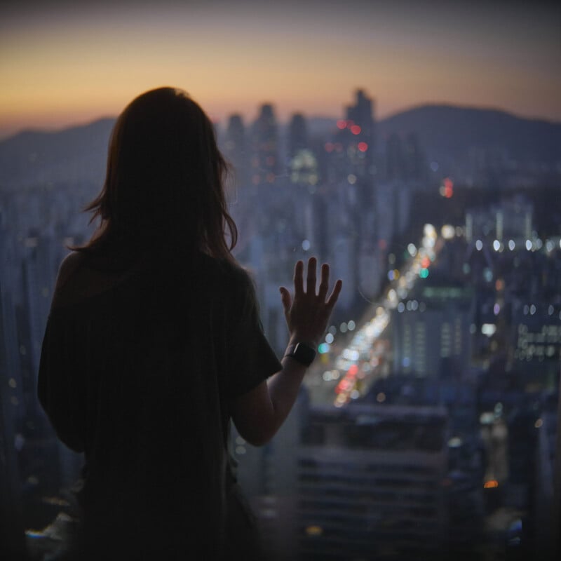 Silhouette of a person with long hair standing at a window, touching the glass, and looking out at a cityscape during twilight. The city lights illuminate the skyline, and the scene conveys a sense of contemplation and tranquility.