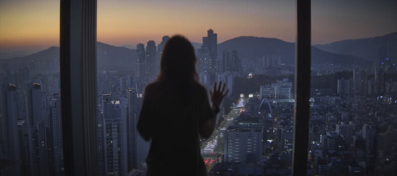 Silhouette of a person standing by a large window, overlooking a cityscape as the sun sets. Skyscrapers and mountains are visible in the distance, while streetlights line a busy road below.