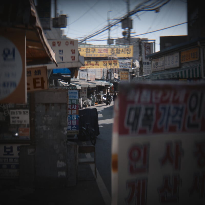 A bustling market street lined with numerous signs in Korean. The narrow road is lively with pedestrians and vendors. Stalls with awnings and various advertisements create a vibrant and busy atmosphere.