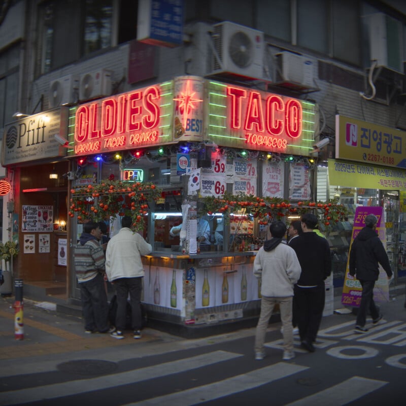 A street food stall named "Oldies Taco," decorated with red neon lights. People are gathered around, ordering food. The stall displays colorful signage and is adorned with flowers. Nearby stores are visible, adding to the lively urban scene.