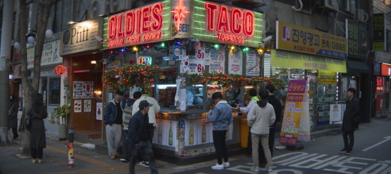 People standing in line at a street food stall called "Oldies Taco" in an urban setting, with bright signage and decorations. The scene is lively, with customers waiting to order and purchase food. Various signs and advertisements are visible nearby.