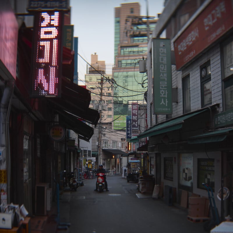 A narrow alleyway in a city lined with various shops and signs in Korean. A person rides a red scooter in the distance, surrounded by tall buildings. Neon lights are visible, adding a vibrant touch to the scene.