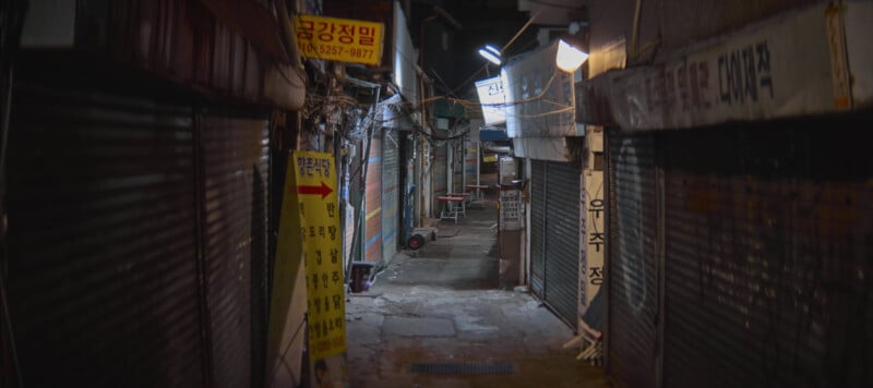 Narrow alleyway at night with closed shop shutters and dim, ambient lighting. Signs with Korean text hang on the walls, and a small table with seats is visible further down the alley. The scene is empty and quiet.