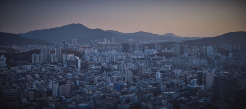 A panoramic view of a sprawling cityscape at dusk, with densely packed high-rise buildings. Rolling hills form the backdrop, and the sky transitions from blue to a warm orange as the sun sets, casting a serene glow over the scene.