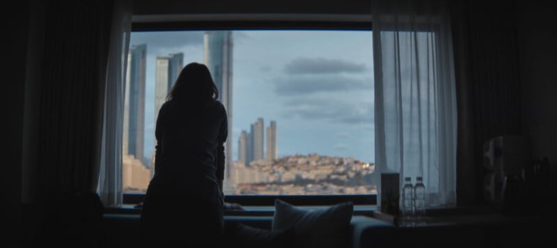 Silhouette of a person leaning on a window ledge, gazing out at a cityscape with tall buildings under a cloudy sky. The scene is framed by curtains, and two water bottles are visible on a table by the window.