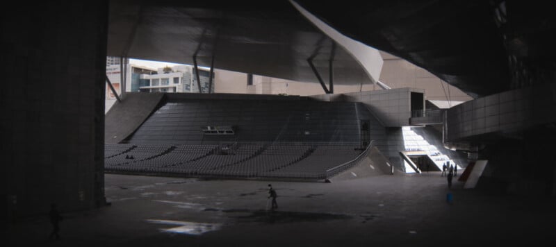 A modern, futuristic building interior with gray geometric structures and seating on one side. There are a few people walking and skateboarding on the wet floor, creating a dynamic scene in the dimly lit area.