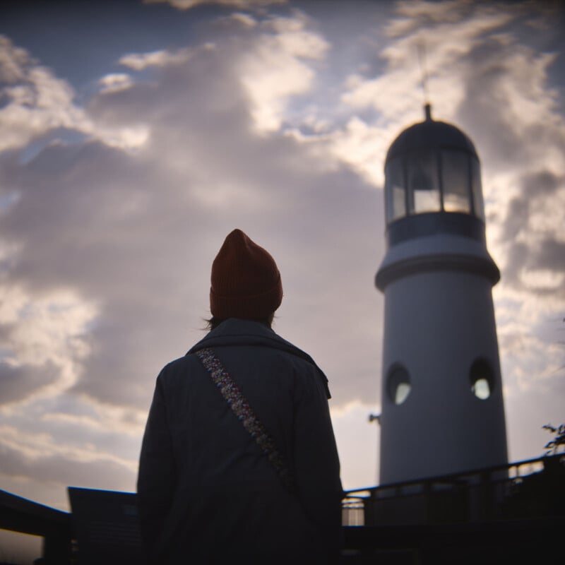 A person wearing a coat and beanie stands in silhouette, facing a tall lighthouse against a backdrop of cloudy sky. The scene conveys a sense of contemplation and tranquility.