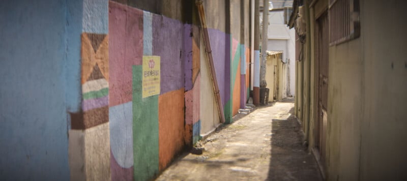 A narrow alley with colorful geometric murals on one side and plain walls on the other. The path is empty and leads to a bright area in the distance.