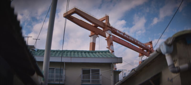 A large orange crane looms over a building with a green roof and balconies. The sky is partly cloudy, and there are several poles and cables in the foreground.