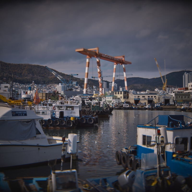 A bustling harbor scene with several docks and moored boats under a cloudy sky. Large orange cranes and industrial buildings are visible in the background, with hills rising behind them.