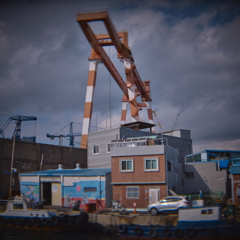 A coastal industrial scene featuring a large, orange-and-white gantry crane towering over buildings. A small boat is docked in the foreground, and the sky is overcast with clouds.