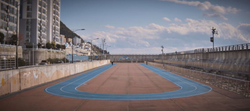 A blue running track with orange lanes is situated between tall buildings on one side and a concrete barrier on the other. The track leads towards a view of distant mountains and a blue sky with scattered clouds.