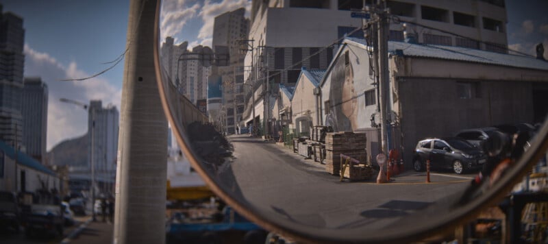 A curved mirror reflects an urban street scene with buildings, parked cars, and a narrow sidewalk. The sky is partly cloudy, and more buildings are visible in the background. The mirror creates a wide-angle effect of the area.