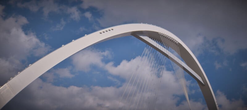 A modern, white arch bridge with suspension cables set against a cloudy blue sky. The angle emphasizes the curve of the arch and the geometric lines of the cables.