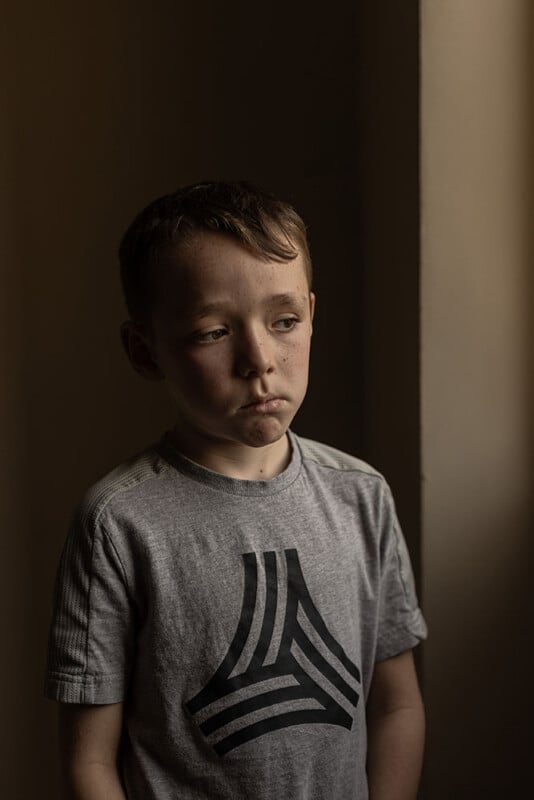 A young boy stands by a window, looking contemplative. He is wearing a gray T-shirt with a geometric design. The lighting casts soft shadows, highlighting his expression as he gazes outside.