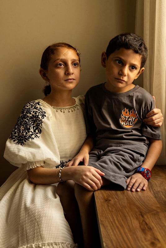 A young girl in a white dress with black embroidery holds a young boy wearing a gray shirt and shorts with "Stay Mad" in flaming letters. They are sitting together on a wooden surface near a window, looking thoughtfully outside.