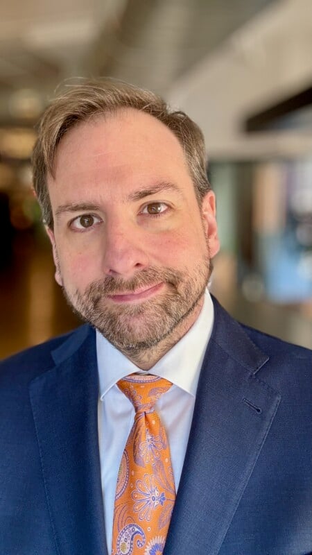 A person with short hair, a beard, and a mustache is wearing a blue suit and an orange paisley tie. The background is blurred, suggesting an indoor setting. The person is smiling gently and looking directly at the camera.