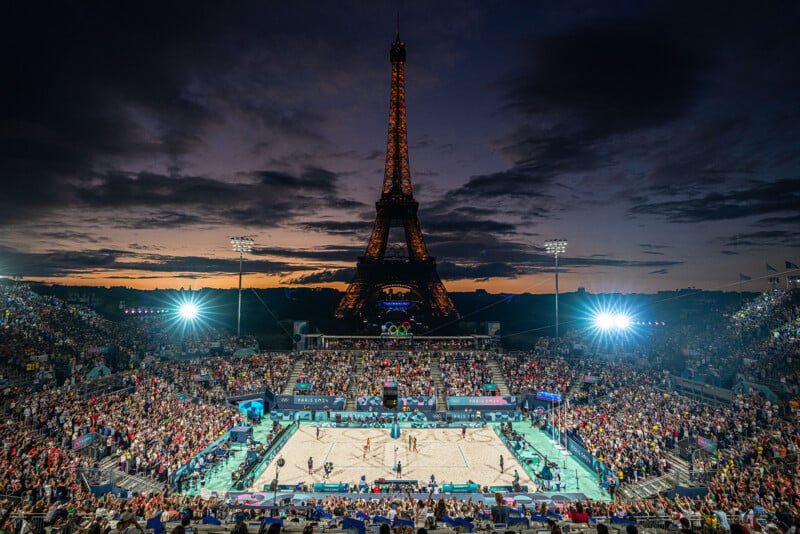 A crowded outdoor volleyball event takes place at night with the Eiffel Tower illuminated in the background. Stadium lights shine brightly on the court as the sky is painted with deep shades of dusk. Spectators fill the stands around the sandy court.