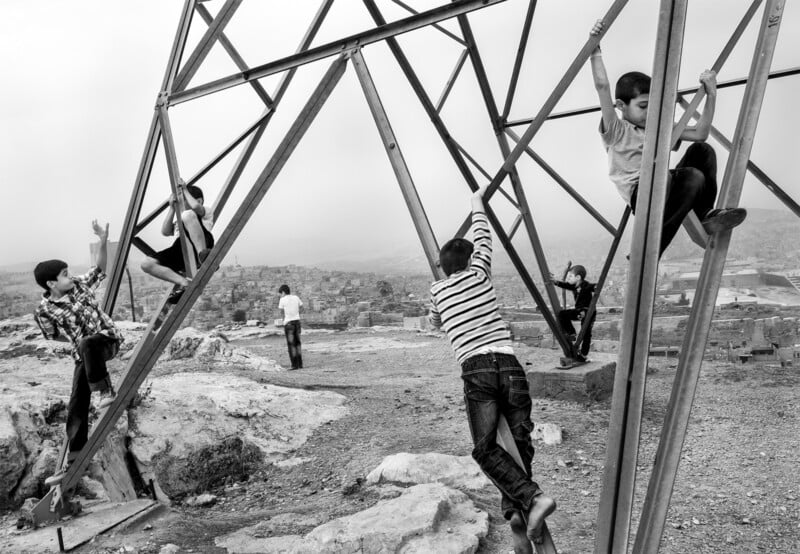 Children climb and play on the metal structure of an electricity pylon in an open area overlooking a cityscape. The black-and-white image captures the energetic scene against a cloudy sky and distant urban background.