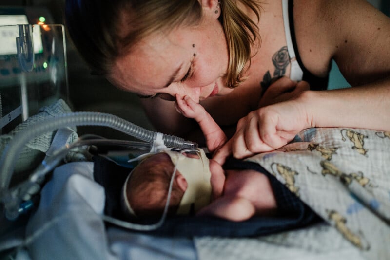 A woman gently touches a newborn's face in an incubator. The baby is wearing a breathing mask and is wrapped in a blanket. The scene is tender and intimate, reflecting a strong bond and care.