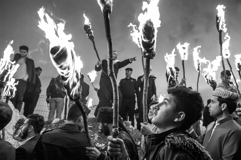 A group of people holding burning torches at night. One man in the foreground looks upward, while others stand in the background. Two individuals, one pointing, appear elevated on a hill, surrounded by a cloudy sky, suggesting a gathering or event.