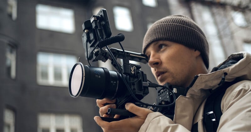 A person in a knit hat and beige jacket holds a professional camera rig, intently focusing on capturing footage. They are standing outdoors with blurred buildings in the background.