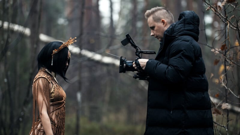 A filmmaker in a black jacket films a woman outdoors in a forest. The woman, wearing a brown, textured outfit and a headpiece, faces the camera. The background is filled with tall trees and dim natural light.