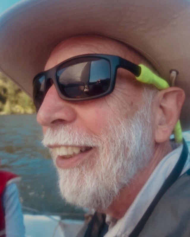 A man with a white beard and sunglasses smiles while wearing a wide-brimmed hat. He is outdoors on a boat, with water and trees visible in the background.