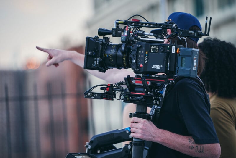 A person operating a professional ARRI film camera on set. They are wearing a cap and pointing into the distance. Another person stands nearby. The setting appears outdoors, with buildings in the background.