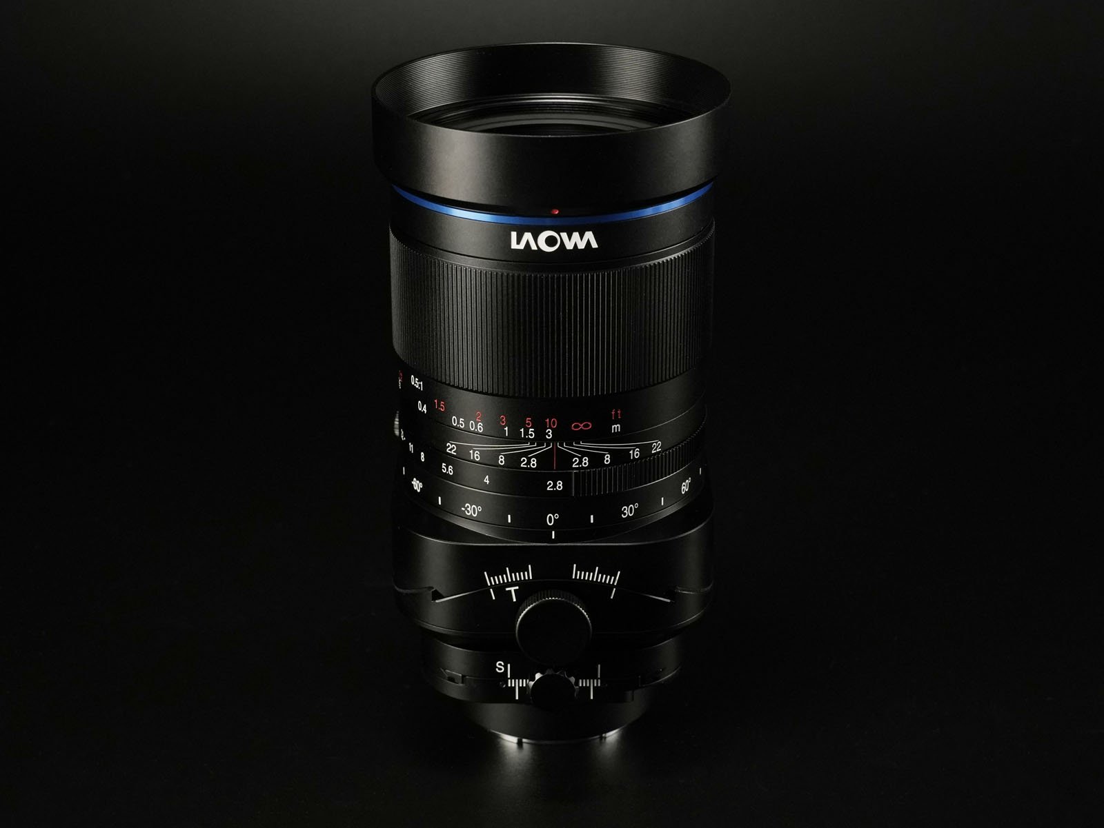 Close-up of a black Laowa camera lens standing vertically against a dark background. The lens has visible focus and aperture rings with white and red markings, and a blue ring near the top.