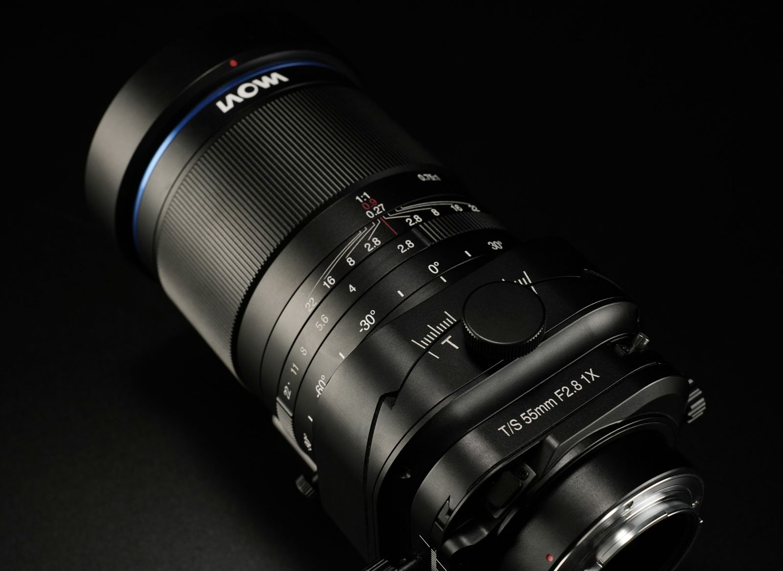 A close-up shot of a black camera lens against a dark background. The lens features various dials and markings, including "T/5 85mm F2.8 1X," with a matte finish and a blue accent near the top.