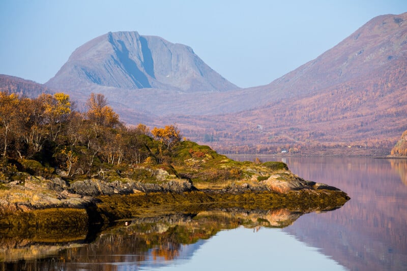 A serene landscape features a calm lake reflecting the surrounding autumn foliage and distant mountains under a clear blue sky. The scene is peaceful, with warm tones of orange and brown dominating the trees and rocky shoreline.