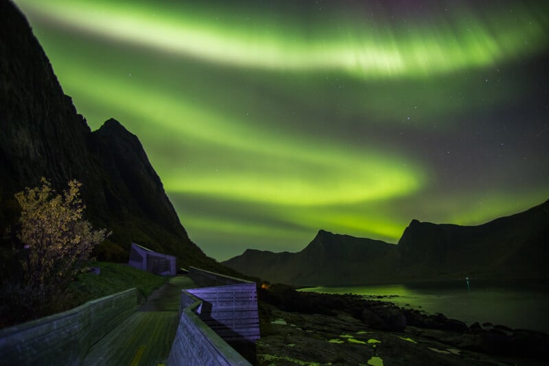 A vibrant display of green Northern Lights illuminates the night sky over a mountainous landscape. The foreground features a wooden path and small structures, with a calm water body reflecting the lights.