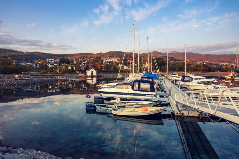 A tranquil marina with several boats docked along a pier. The water reflects the clear sky and surrounding hills. In the background, a small town with colorful autumn trees is visible, set against rolling hills.