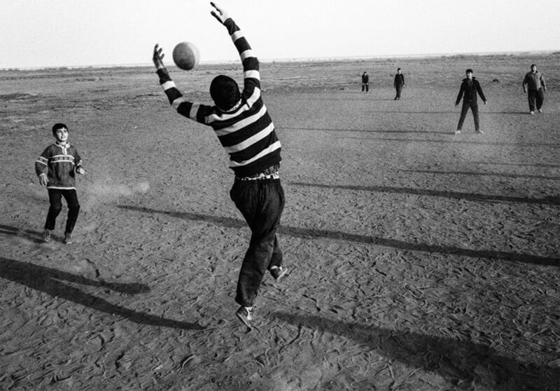 A group of people play soccer on a barren, dirt field. One person in the foreground leaps to catch a ball in mid-air, while others stand scattered in the background. The scene is captured in black and white, emphasizing movement and shadow.