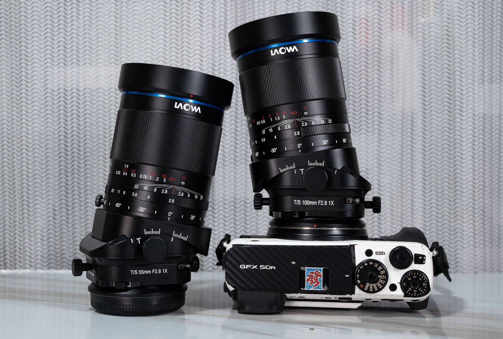 Two Laowa camera lenses are positioned upright on a surface next to a GFX 50R camera. The lenses are labeled "TS 50mm F2.8 1X" and "TS 110mm F2.8 1X." A textured gray background is visible.