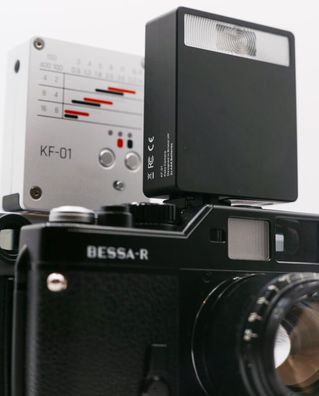 Close-up of a black Bessa-R film camera with a lens attached. On top, there's a silver KF-01 light meter and a black electronic flash unit. The camera is angled slightly upwards, and the focus is on its vintage design features.