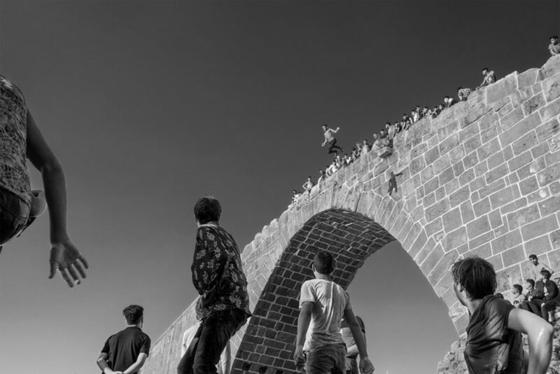 A group of people gathers on and below a large stone bridge. One person is mid-air, jumping off the bridge. The sky is clear and the image is in black and white. People are watching the jumper from different angles.