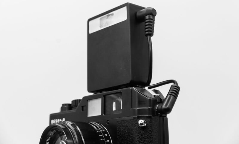 Close-up of a vintage camera with a mounted external flash. The camera has a textured body and a detailed lens, with a cable connecting the flash unit on top. The background is plain and light-colored.