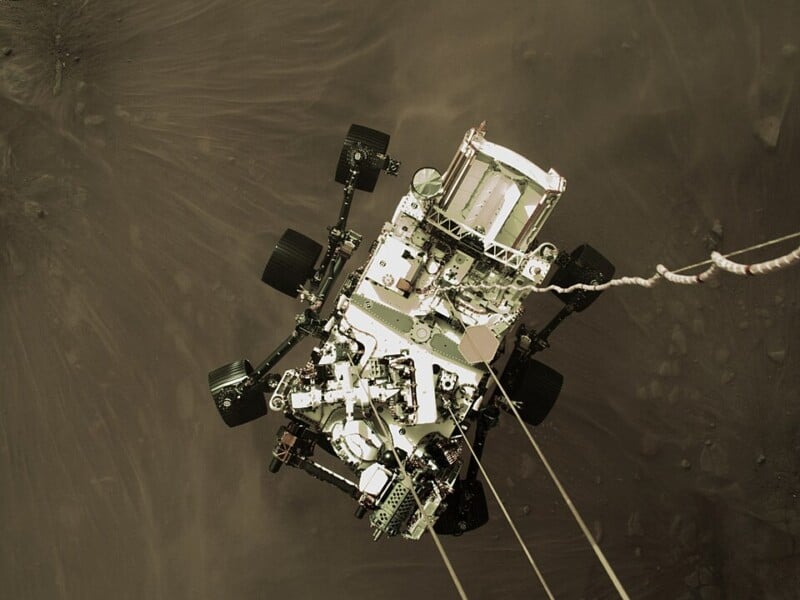 A spacecraft's rover is being lowered onto a dusty, rocky surface with visible cables. The rover is equipped with wheels and various scientific instruments. The background is a textured terrain, hinting at a planetary landscape.