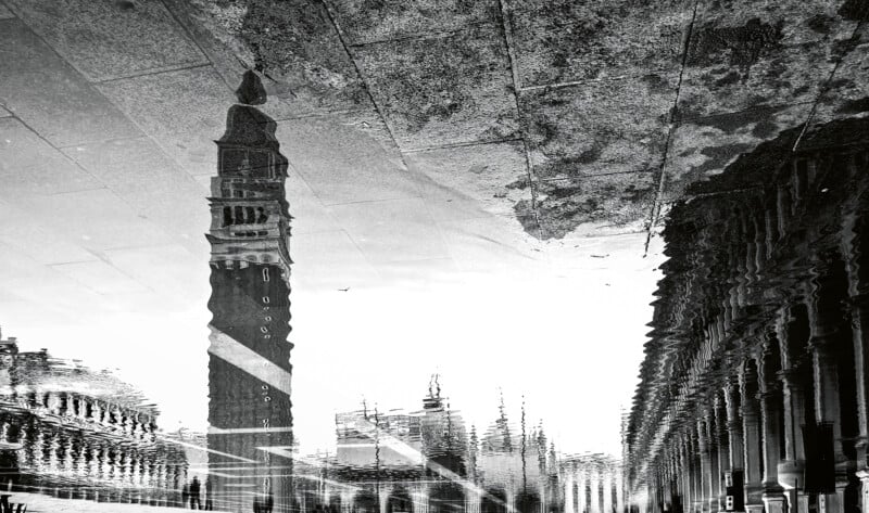 A black and white reflection of buildings in a puddle, with a tall clock tower and a row of ornate columns. The water's ripples create a distorted, abstract effect.