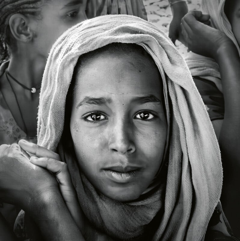 A black and white portrait of a young person with an intense gaze, wearing a headscarf. Their hands are gently resting near their face. The background is slightly blurred with other figures partially visible.