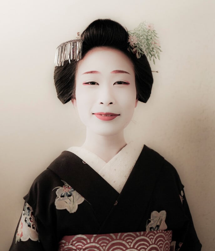 A person wearing traditional Japanese attire, including a black kimono with floral patterns, white undergarment, and red obi. The hairstyle is adorned with decorative hair ornaments. The face is painted in traditional white makeup.